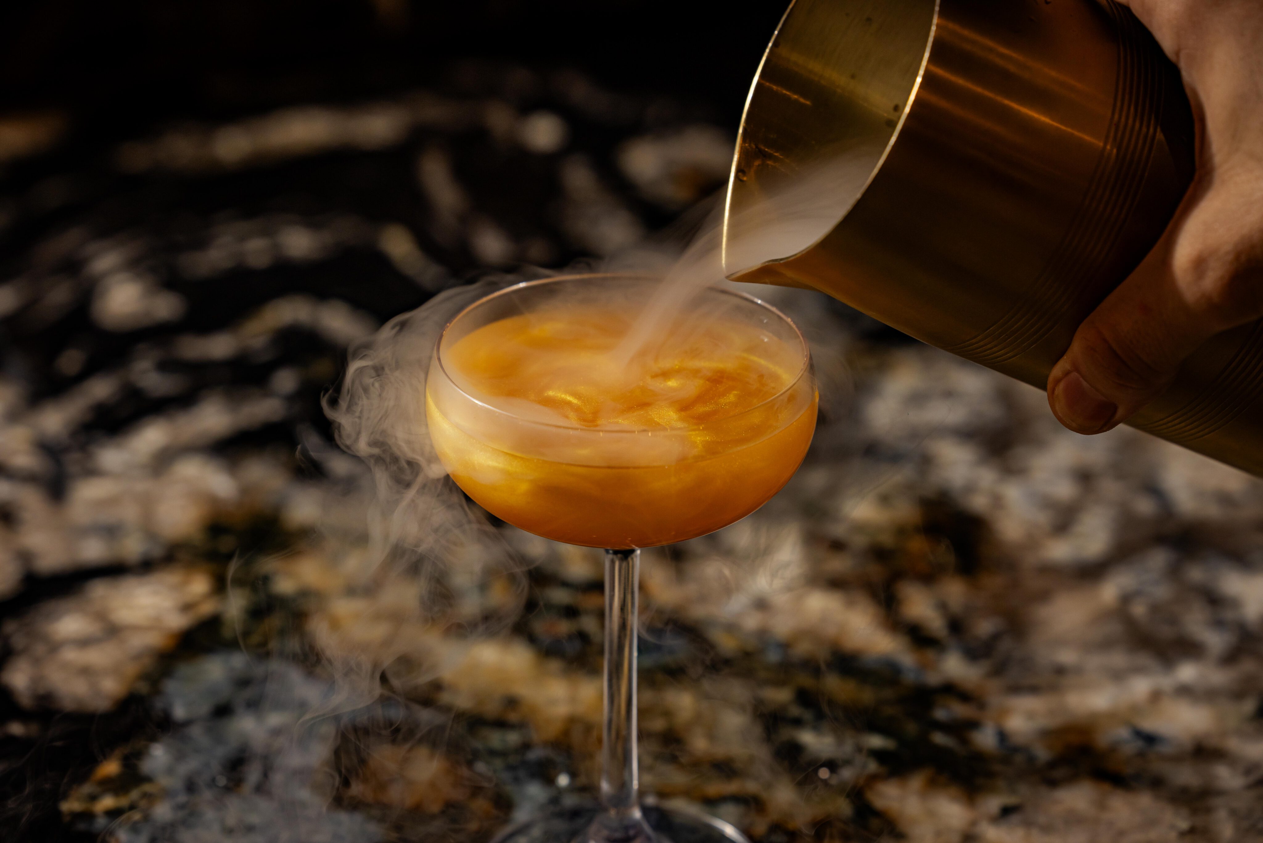 A golden cocktail in a coupe glass is being poured from a shaker, releasing vapor or smoke, on a dark marble surface—an elegant scene perfect for Private Events.