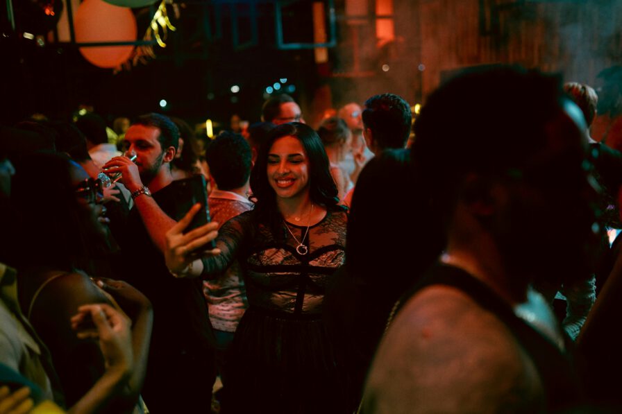 A woman in a black dress smiles while taking a selfie in a crowded, dimly lit party or club, surrounded by people socializing.