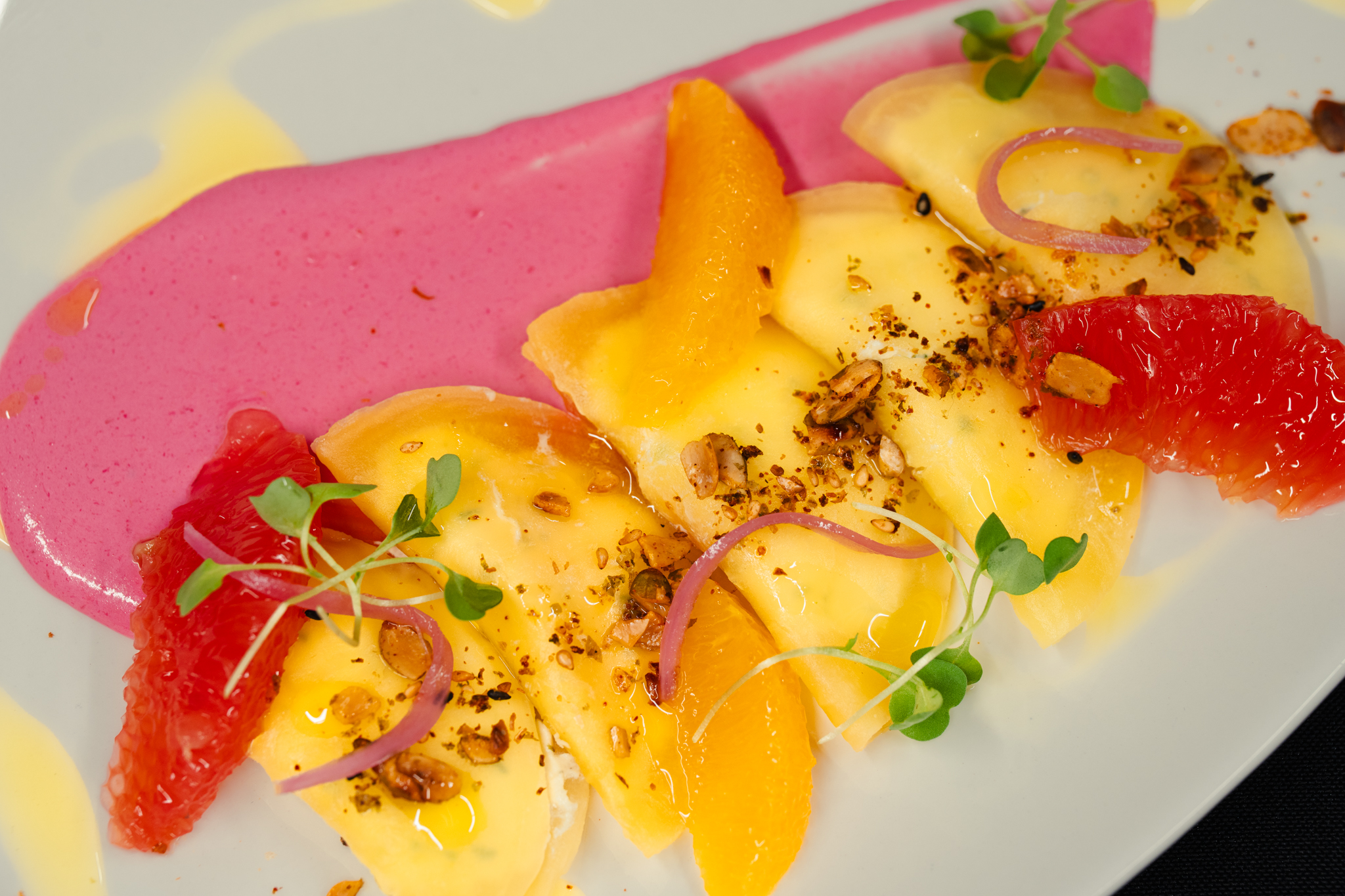 Five ravioli on a pink sauce, garnished with orange and grapefruit segments, microgreens, red onion slices, and crushed nuts on a white plate—perfect for a memorable dinner or as a standout feature on your Dessert Menu.
