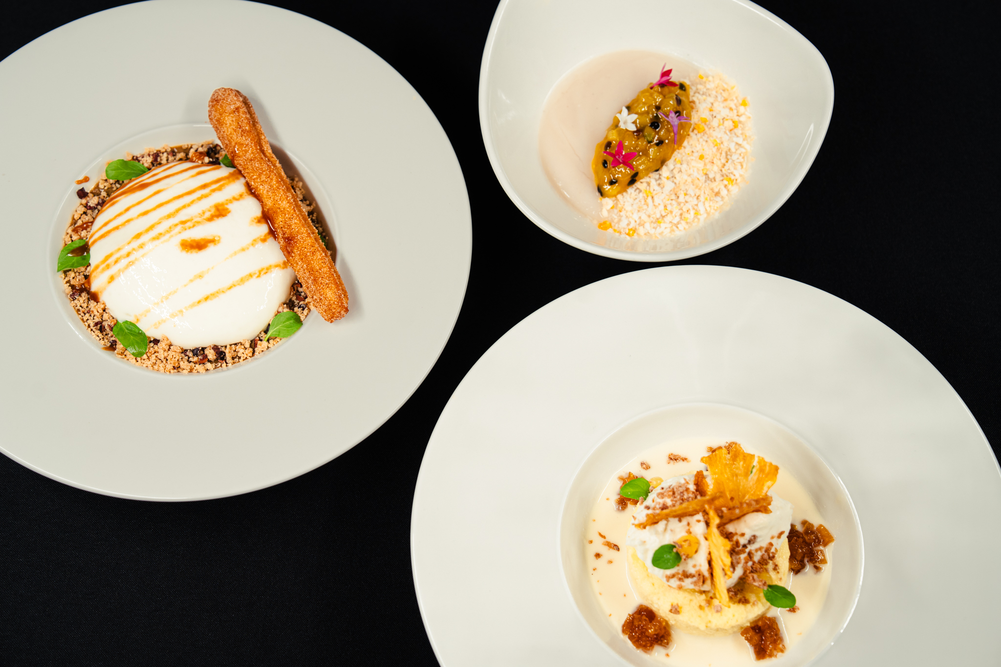 Three white plates on a black surface, each featuring a different plated dessert from the dinner menu, showcase an artful arrangement of creams, crumbles, sauce, and garnishes such as herbs, edible flowers, and a churro stick.