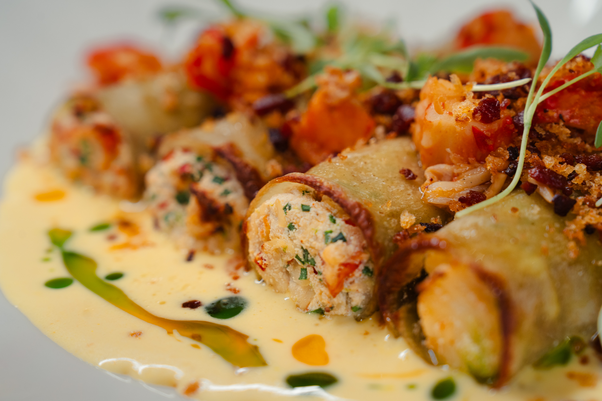 Close-up of rolled pasta filled with a savory mixture, topped with herbs and sauce, and garnished with microgreens and crumbs on a creamy yellow sauce—a perfect dinner option that adds elegance to any menu.