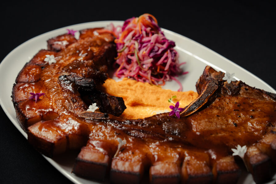 Sliced glazed pork ribs arranged on a white plate with a side of purple cabbage slaw and a serving of orange purée, garnished with small edible flowers—perfect for an elegant dinner presentation or as a savory highlight before exploring the dessert menu.