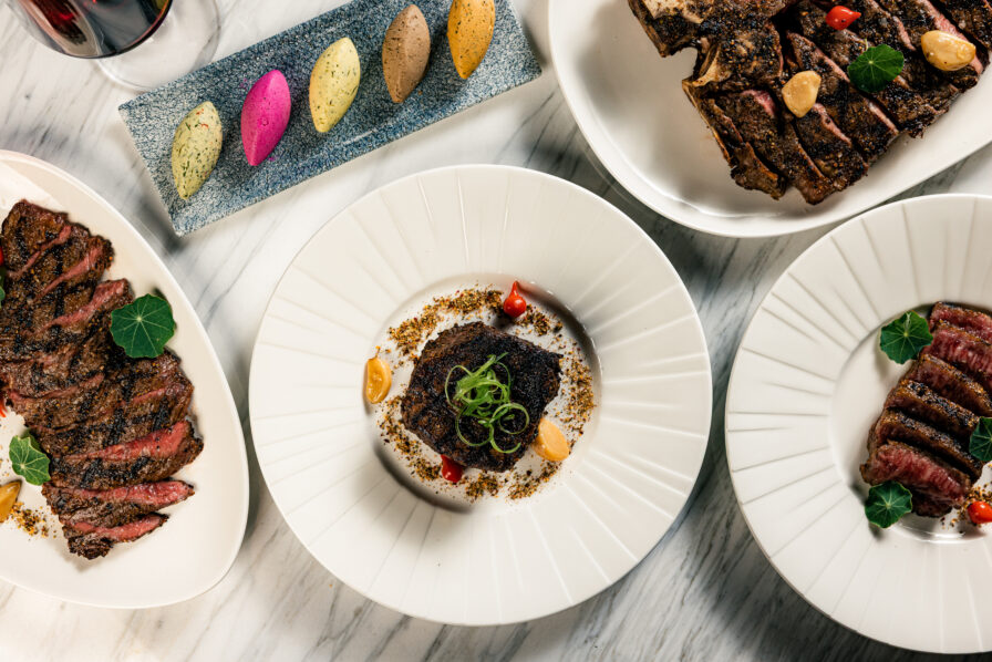 Four plates with sliced steak, sauces, and garnishes on a marble table, accompanied by a glass of red wine and a rectangular dish holding five colorful dollops of flavored butter.