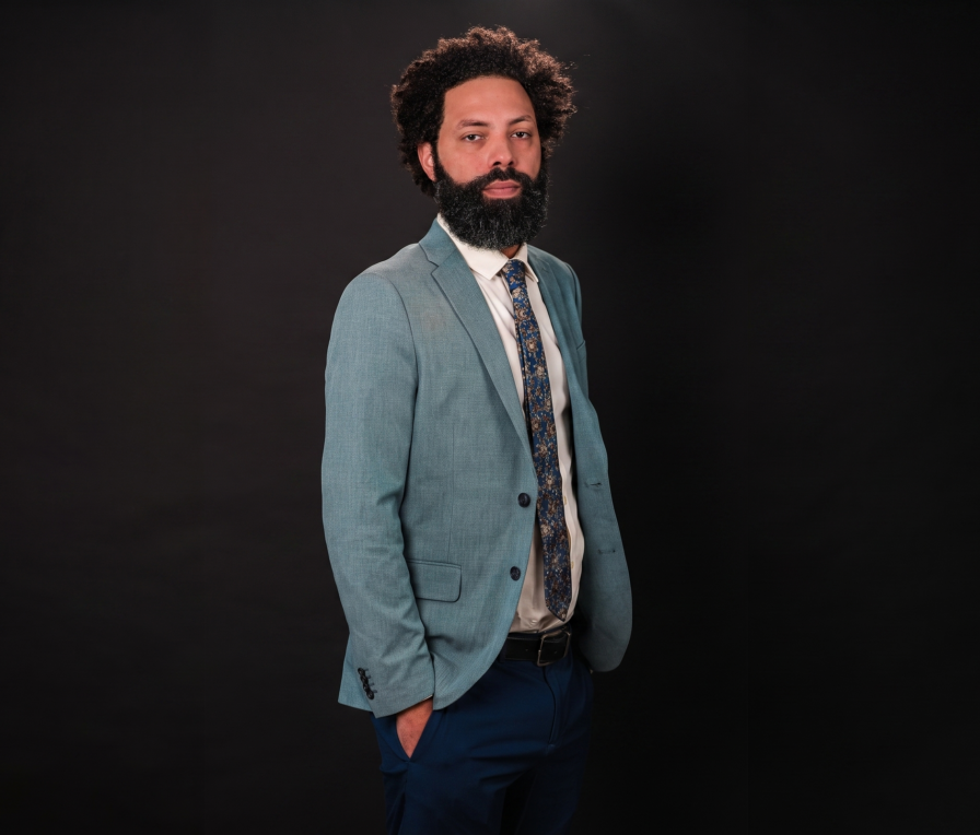 Jesús Bonilla Cepeda, a man with a beard and curly hair, wears a light blue suit jacket, printed tie, and dark pants, standing against a plain dark background with one hand in his pocket.