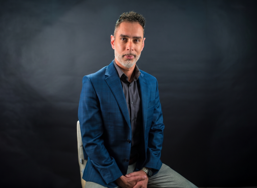 Yuri Oliveira, a man with short dark hair and a trimmed beard, wears a blue blazer and gray shirt as he sits on a chair against a dark studio background, facing the camera with a neutral expression.