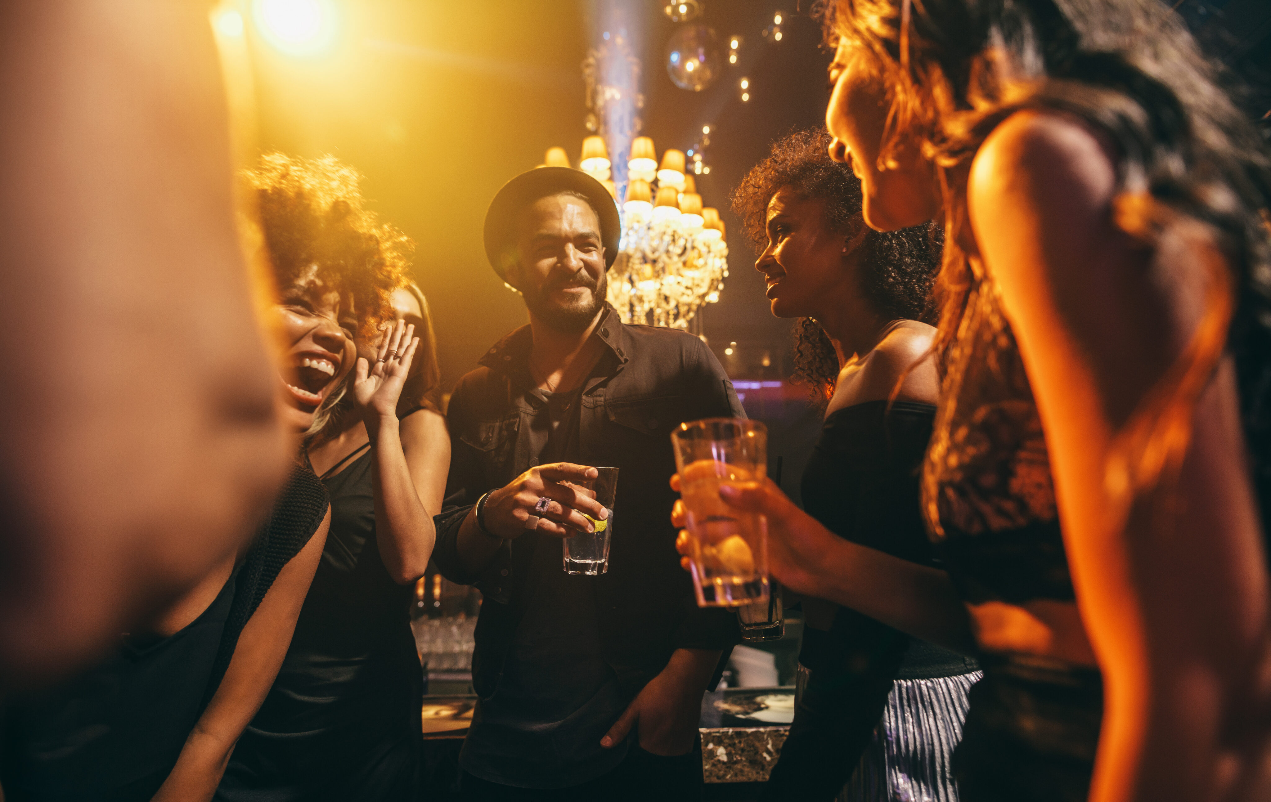 Group of people stand together in a dimly lit bar, holding drinks and talking, with a chandelier visible in the background.