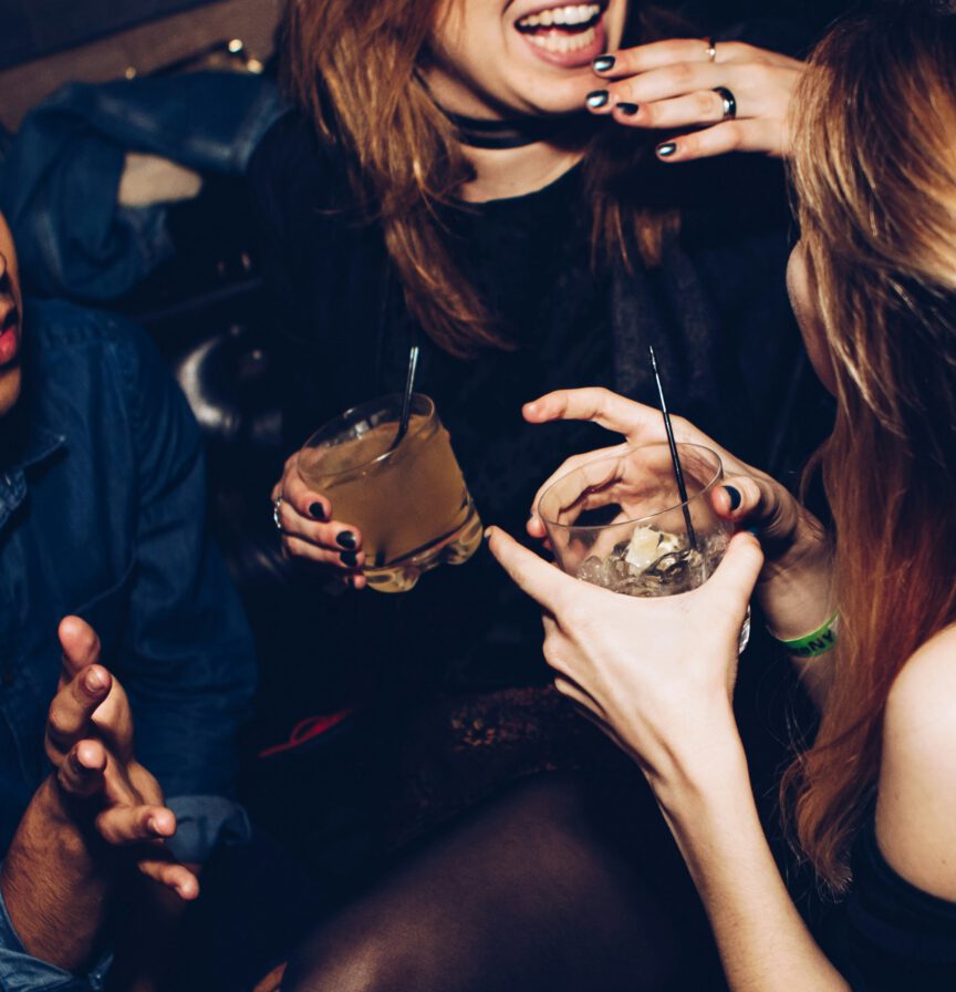 Three people are sitting close together, holding drinks and conversing in a dimly lit setting. Only their hands and parts of their faces are visible.