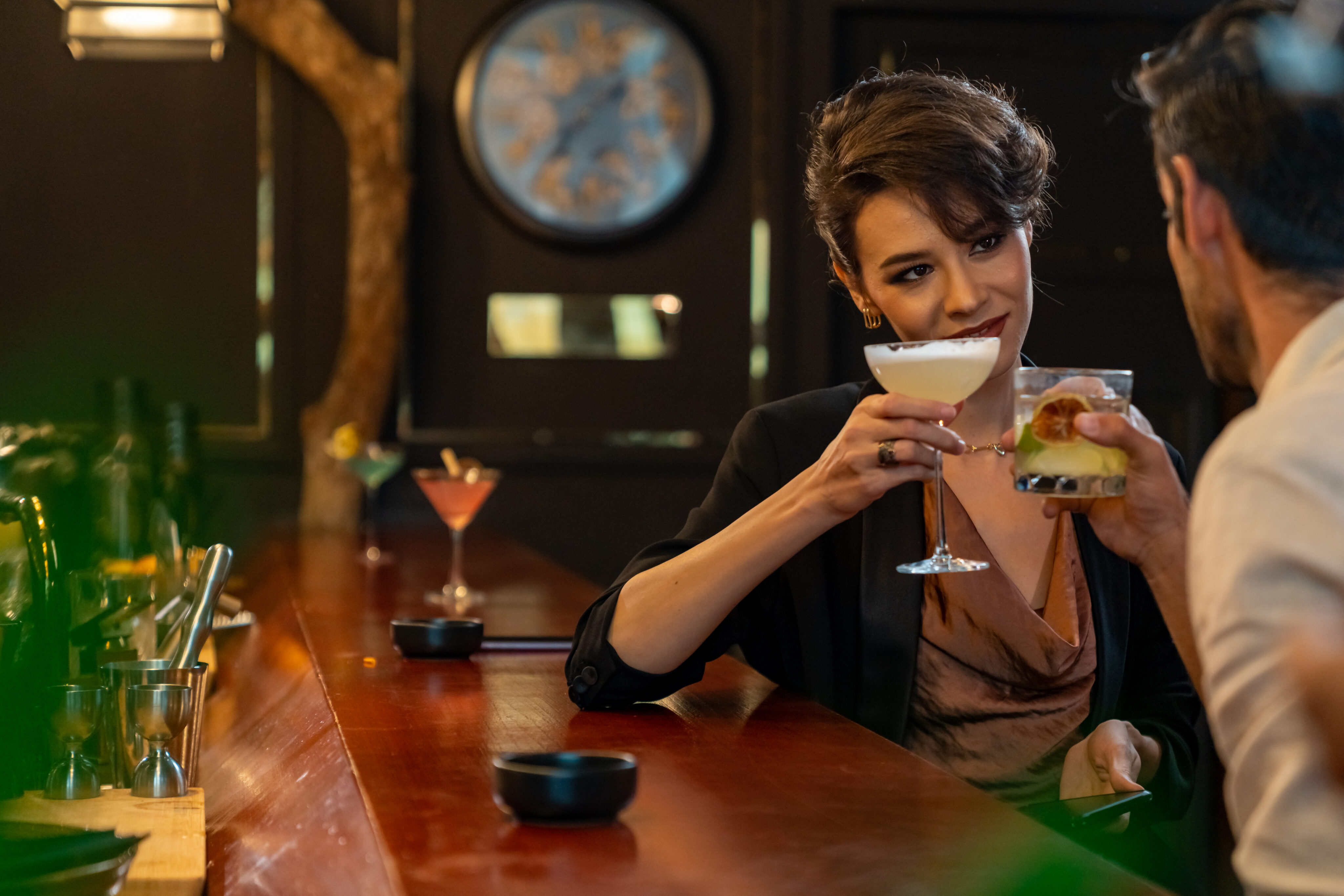 Two people sit at a bar, holding cocktails and facing each other, with a clock and additional drinks visible in the background.