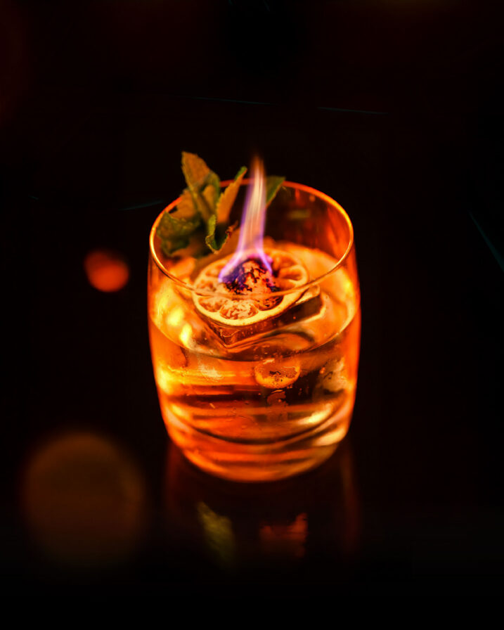 A cocktail in a glass with ice, garnished with a flaming citrus slice and mint, set against a dark background.