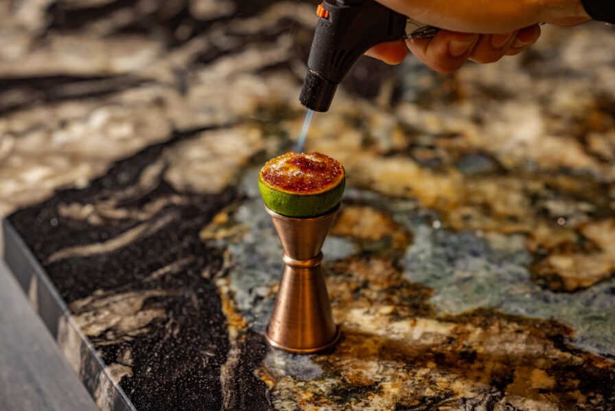 A hand uses a small kitchen torch to caramelize sugar on a halved lime set on a jigger on a marbled countertop.