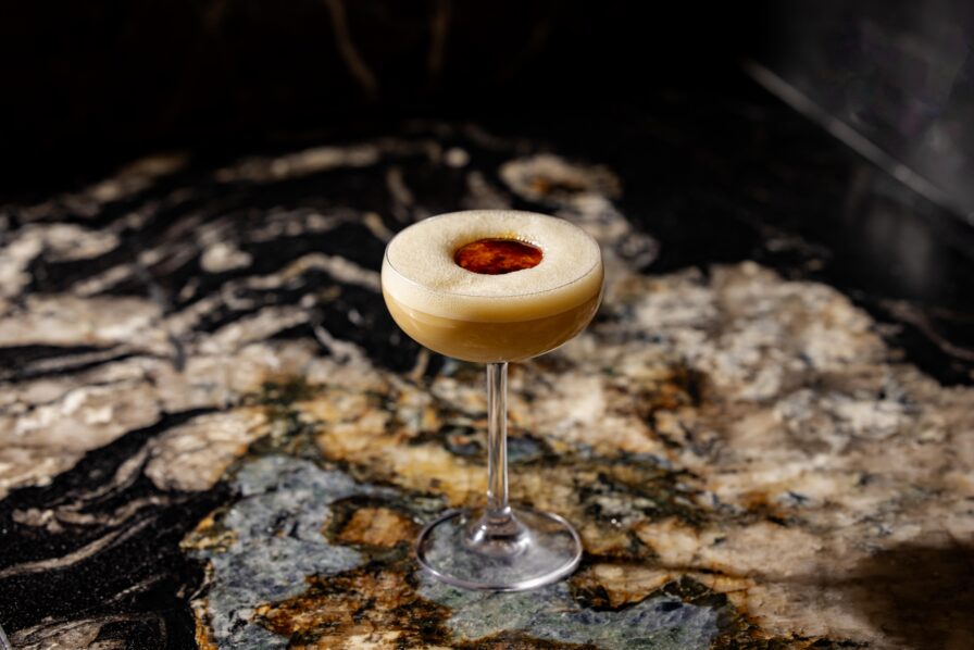 A coupe glass filled with a frothy tan cocktail, garnished with a red circular topping, sits on a polished, marbled stone surface.