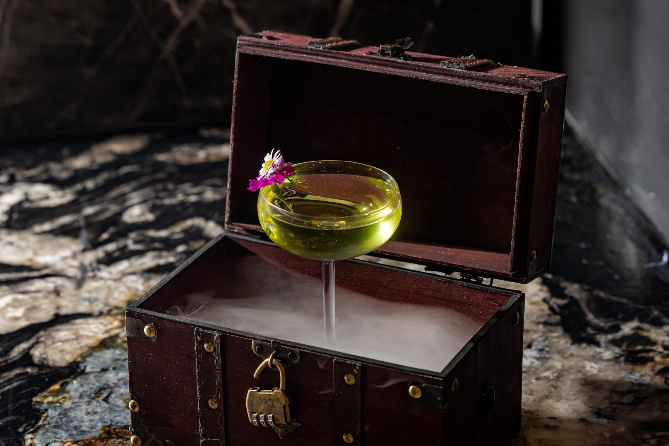 A yellow cocktail in a coupe glass with flowers floats above dry ice inside an open wooden chest, creating a smoky effect on a marble surface.