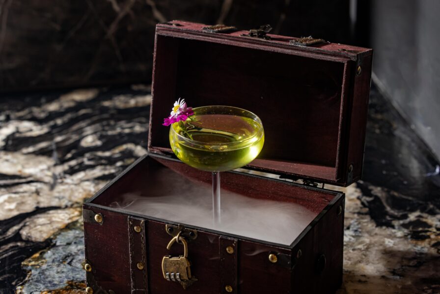 A yellow cocktail in a coupe glass with flowers floats above dry ice inside an open wooden chest, creating a smoky effect on a marble surface.