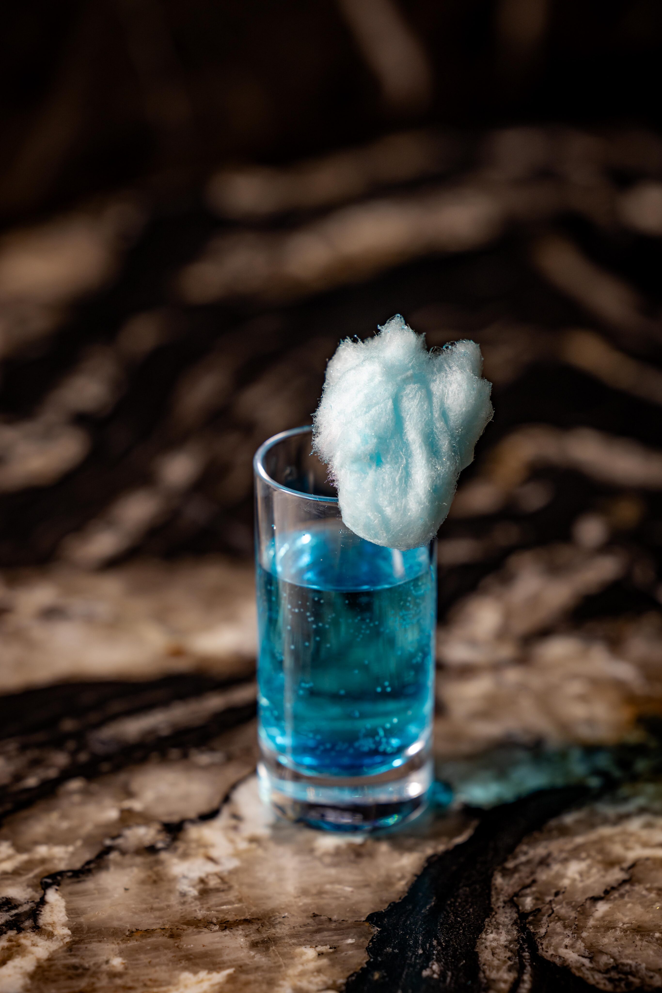 A clear glass filled with blue liquid, topped with a piece of blue cotton candy, sits on a marbled surface.