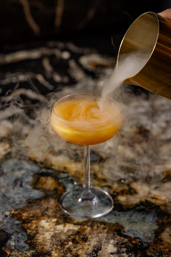 A cocktail glass with an orange drink sits on a marble surface as vapor is poured into it from a metal container, creating a swirling, smoky effect.