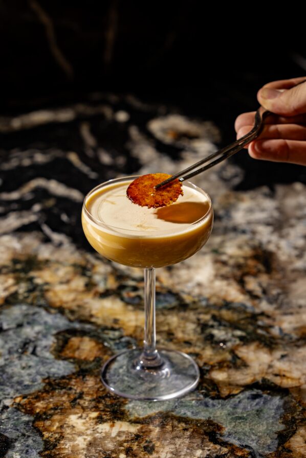 A hand uses tongs to place a thin, round garnish on a creamy cocktail in a coupe glass, set on a marbled surface.