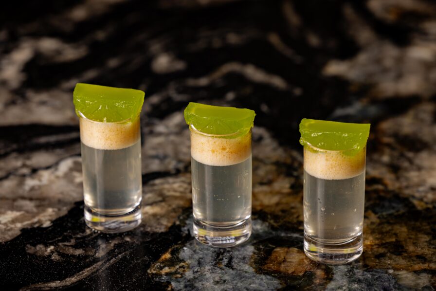 Three shot glasses filled with clear liquid, topped with a green jelly garnish, are placed in a row on a dark, marbled surface.