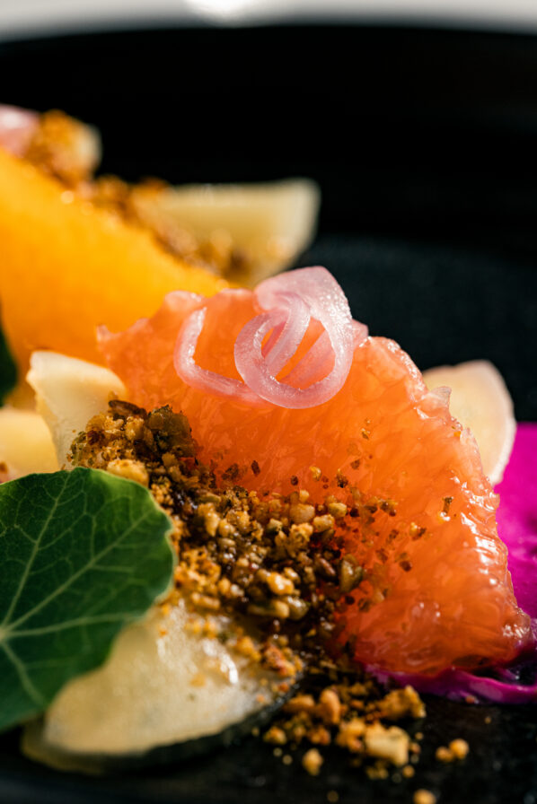 Close-up of a plated dish featuring a pink grapefruit segment, pickled onions, crushed nuts, green leaf, and light yellow garnish on a black plate.