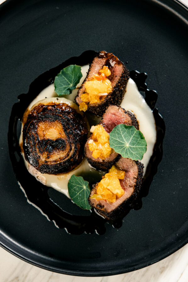Sliced seared meat with orange relish, served on a creamy white puree, garnished with green leaves on a black plate.