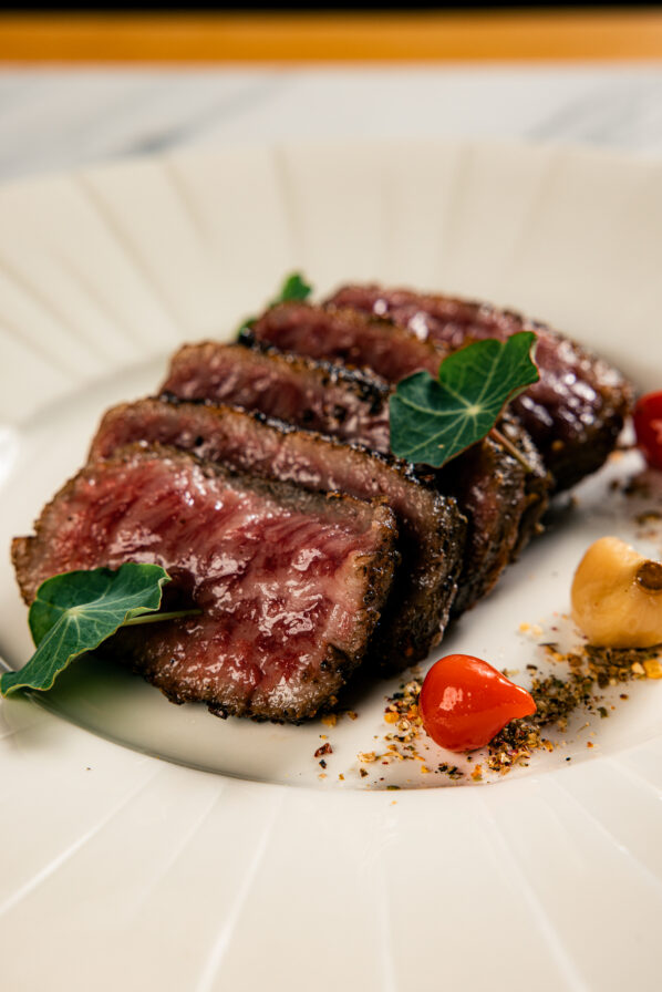 Sliced rare steak garnished with green leaves, small red peppers, garlic cloves, and seasoning on a white plate.