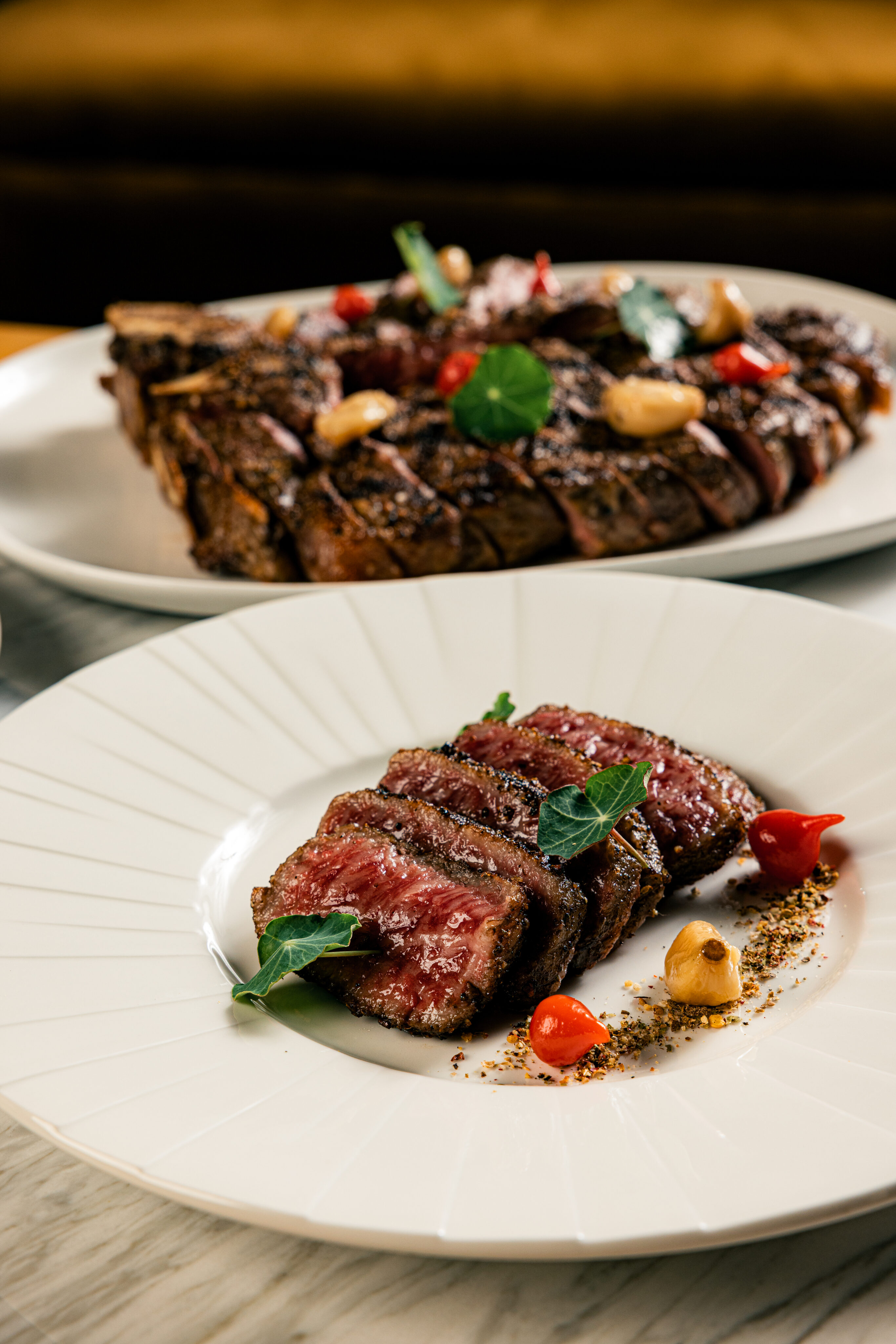 Sliced steak served on a white plate with garnish, roasted garlic, and small red peppers. A larger portion of steak is visible on another plate in the background.