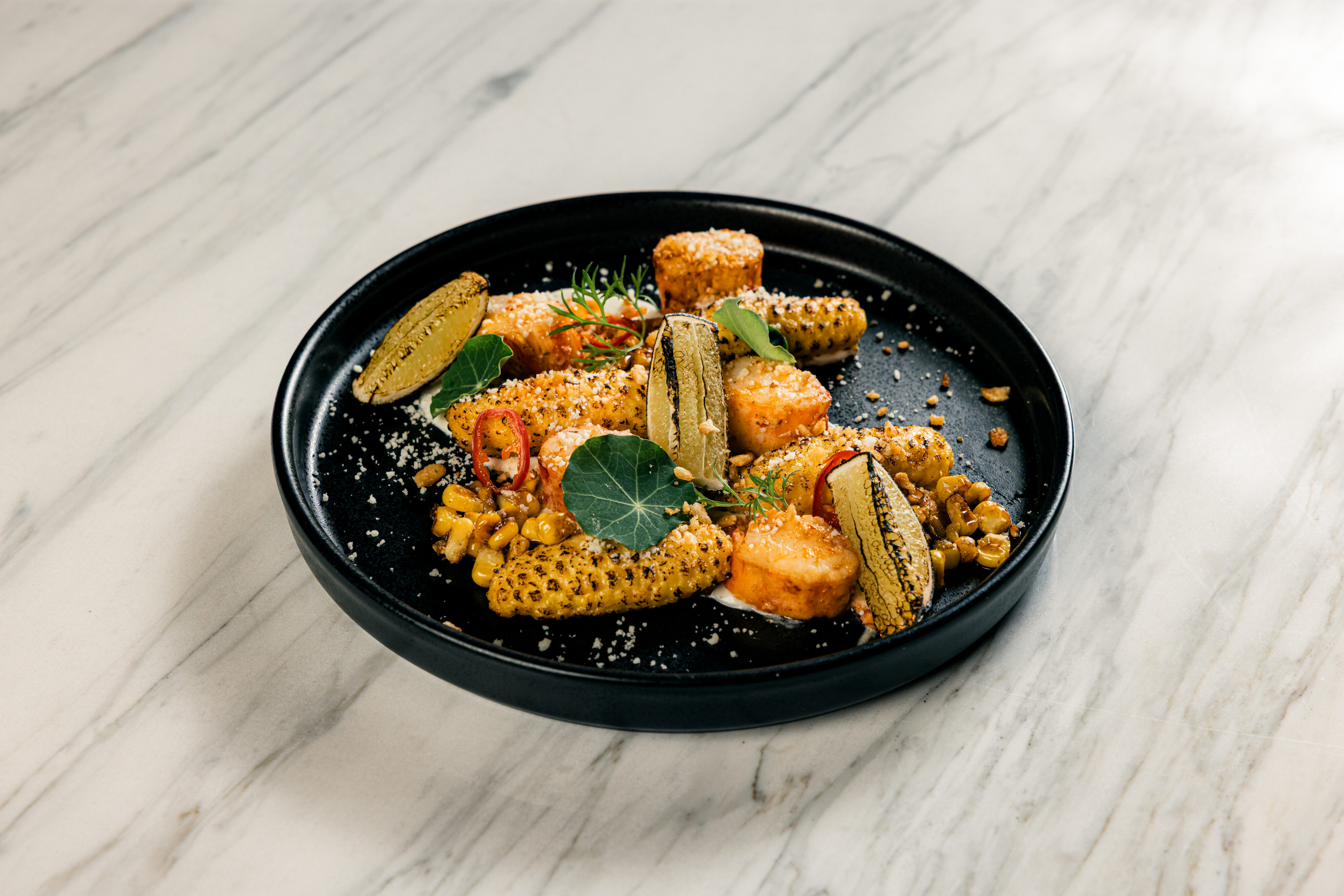 A black plate with grilled corn, cubes of seared cheese, crushed nuts, microgreens, and sauce, arranged on a white marble surface.