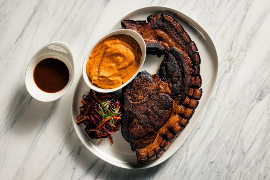 A plate with a large grilled steak, mashed sweet potatoes, a small serving of slaw, and a side of brown sauce, all on a white marble surface.
