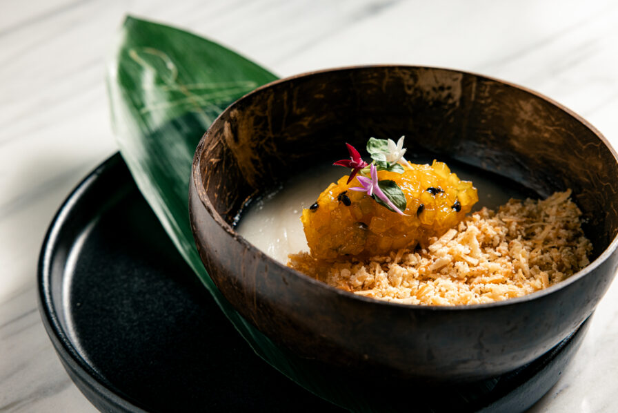A highlight from our Dessert Menu, this wooden bowl features a creamy white dessert topped with yellow fruit, edible flowers, and a crumbled garnish, all elegantly presented on a plate with a green leaf—perfect for ending your Dinner on a sweet note.