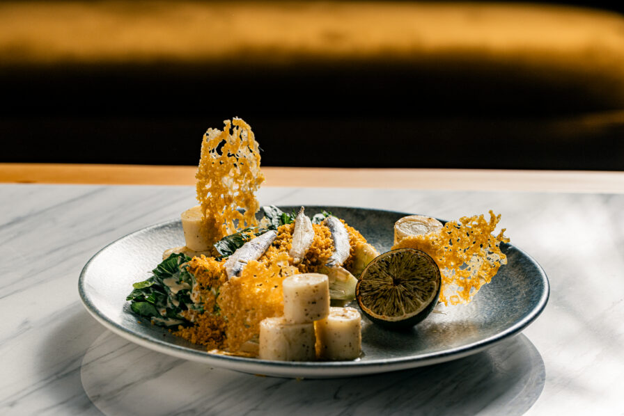A plated gourmet dish perfect for a refined Dinner features cylindrical pieces, leafy greens, crispy cheese garnishes, and a halved grilled lime, elegantly presented on a marble table.