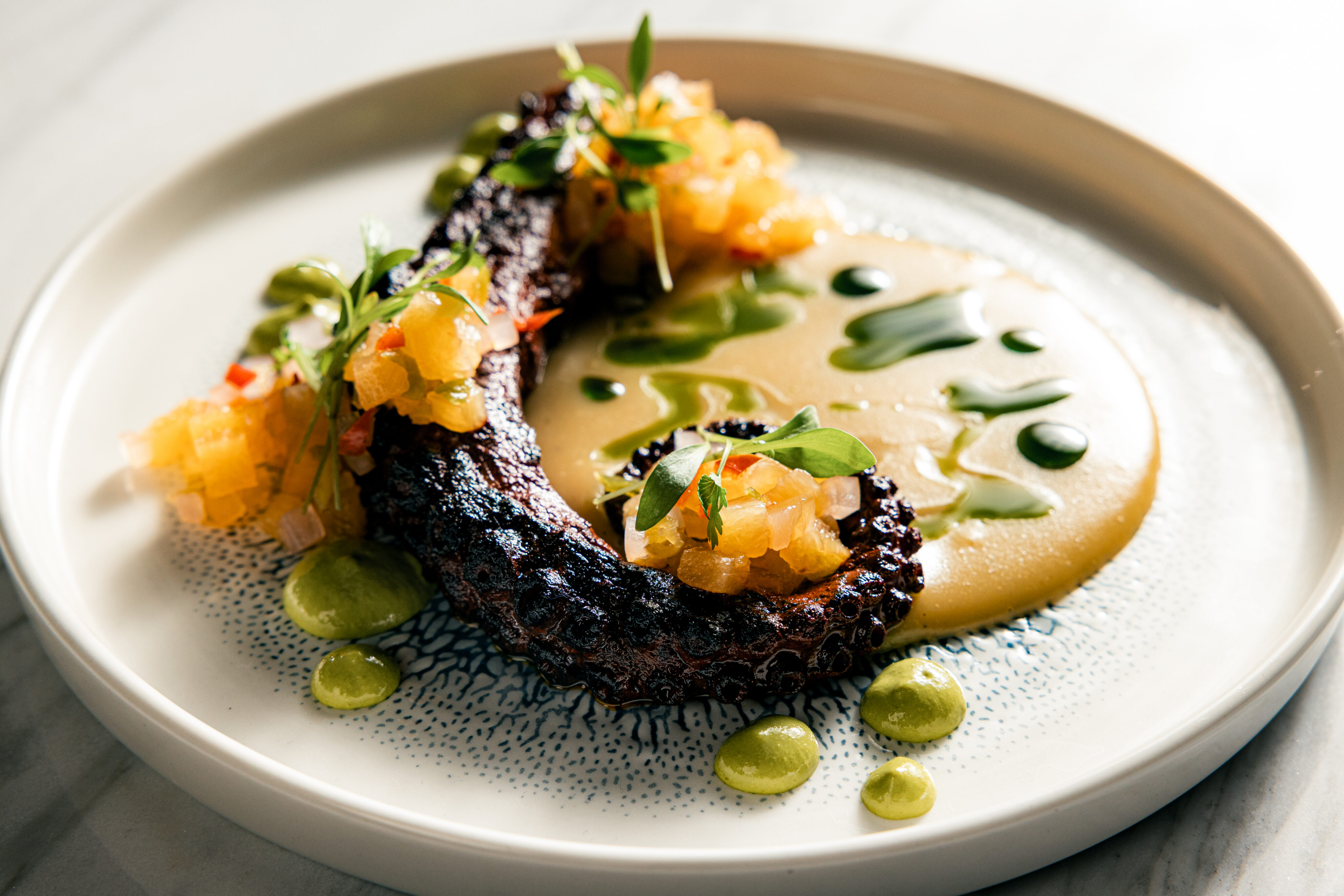 Grilled octopus tentacle with diced garnish on creamy puree, plated with green sauce and microgreens on a white plate—perfect as an elegant dinner highlight before exploring our decadent dessert menu.