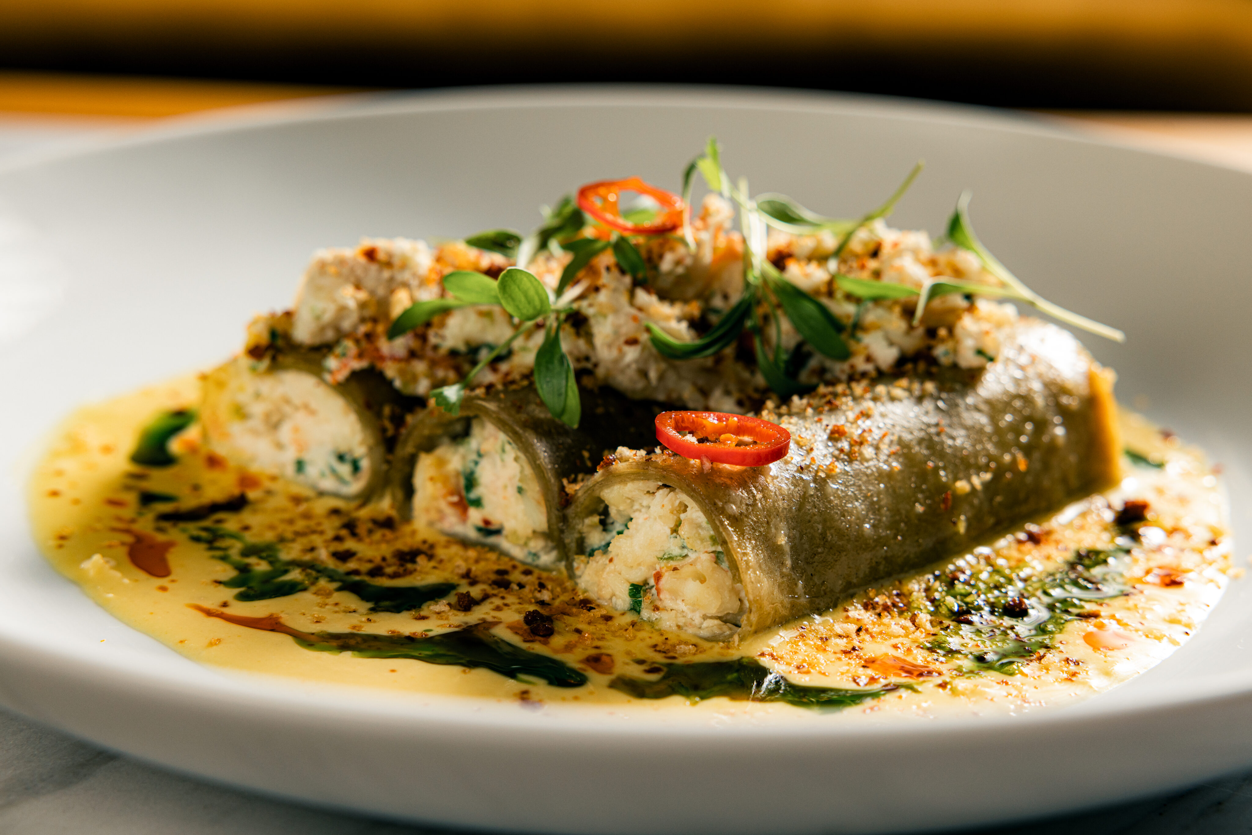Two stuffed pasta rolls topped with ricotta, microgreens, and sliced red chili, served on a sauced white plate with oil and herb garnishes—a delicious choice for your dinner menu.