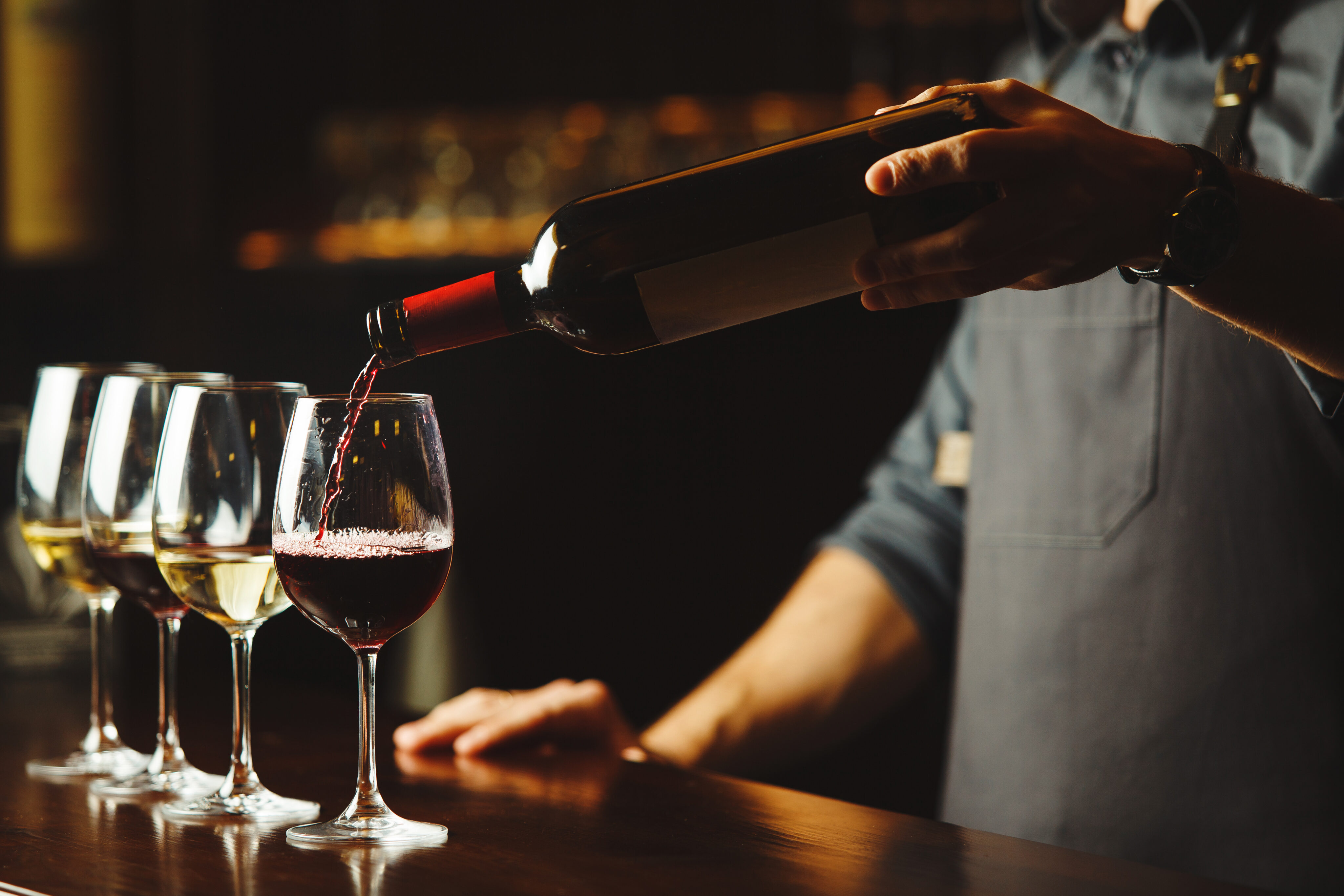 A person pours red wine from a bottle into a wine glass next to two other filled wine glasses on a wooden bar counter.