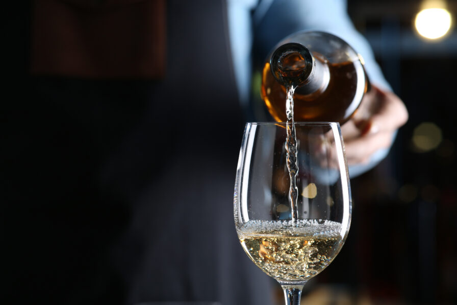 Person pouring white wine from a bottle into a clear wine glass, with some bubbles forming at the top of the glass.