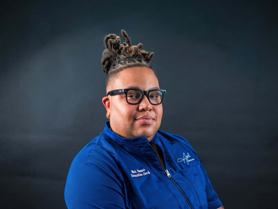 A person with dreadlocks, glasses, and visible tattoos sits on a chair, wearing a blue collared shirt with embroidered text, against a dark background.
