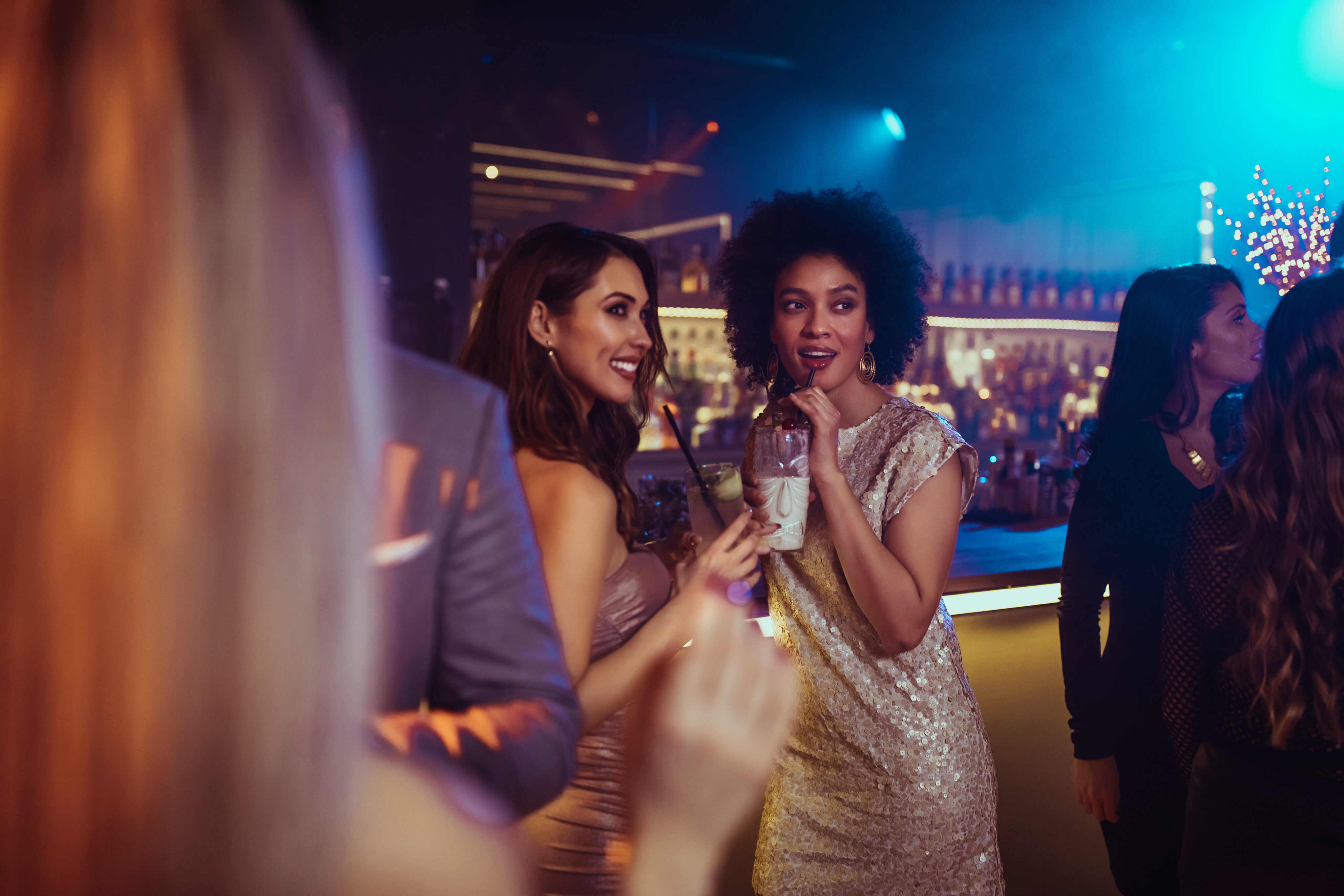 Two women in evening dresses stand together at a bar, one holding a drink, surrounded by other people in a dimly lit nightclub or party setting.