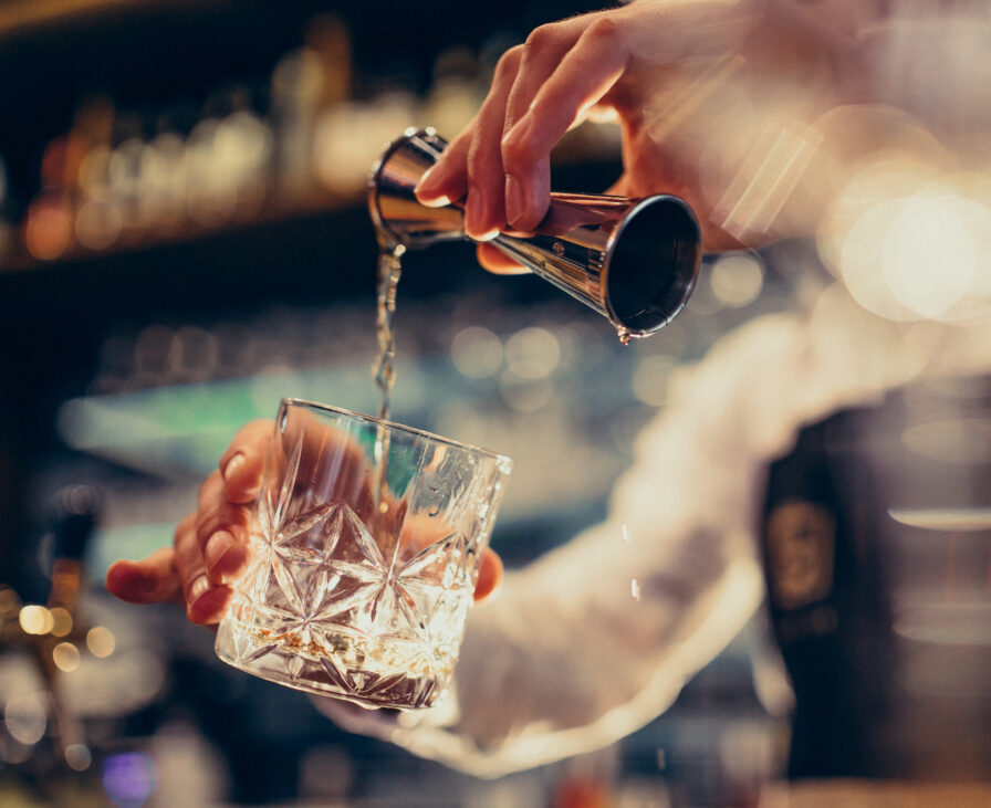 A bartender pours a clear liquid from a jigger into a faceted glass at a bar, with bottles blurred in the background.