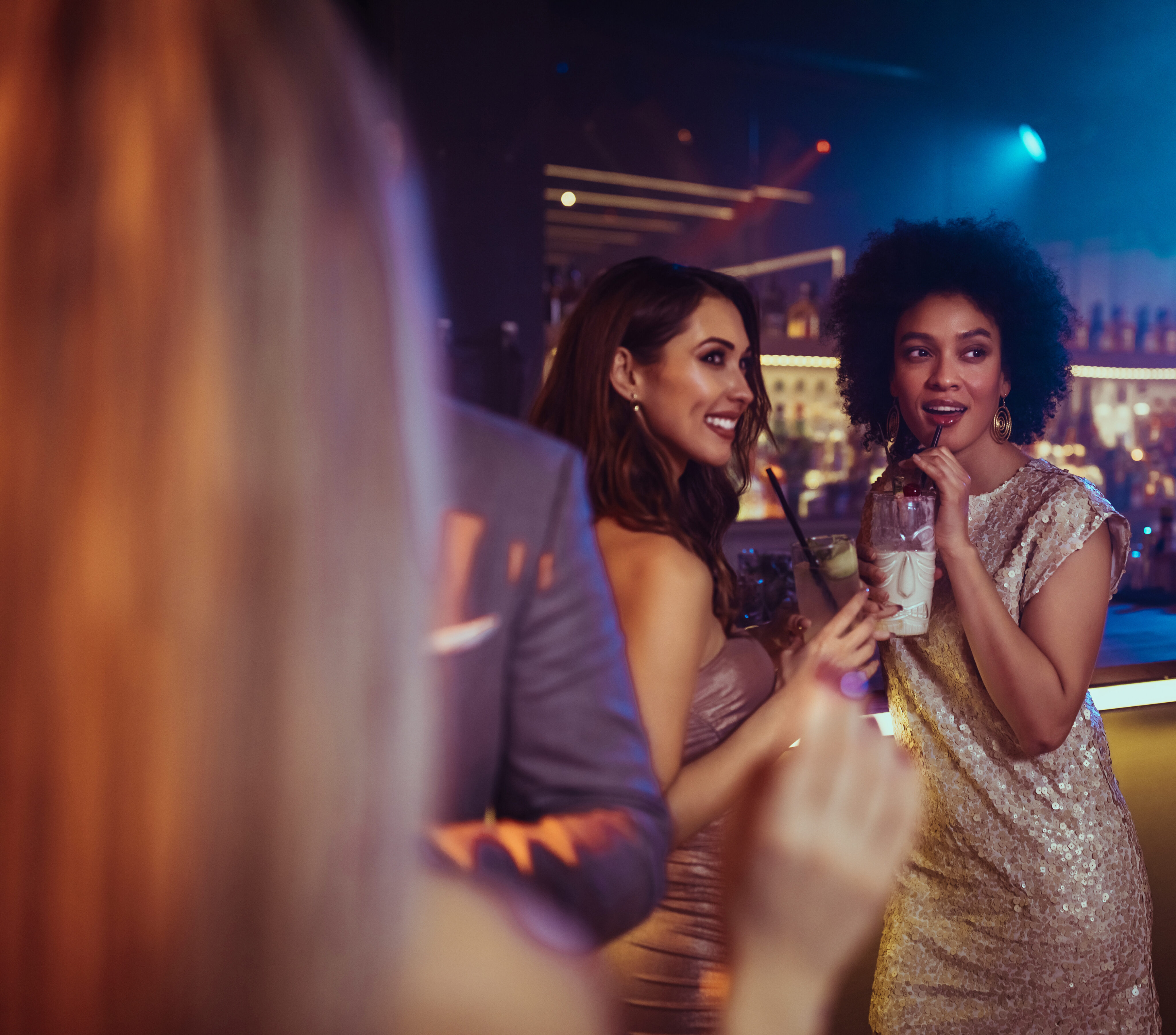 Two women in sparkly dresses stand at a bar with drinks in hand, smiling and talking, while other people socialize in the background.