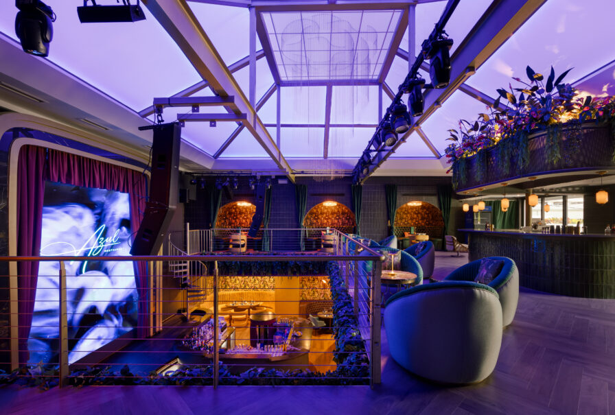 Modern lounge with a stage, neon “Aqua” sign, plush seating, balcony area, skylight ceiling, and ambient purple lighting.