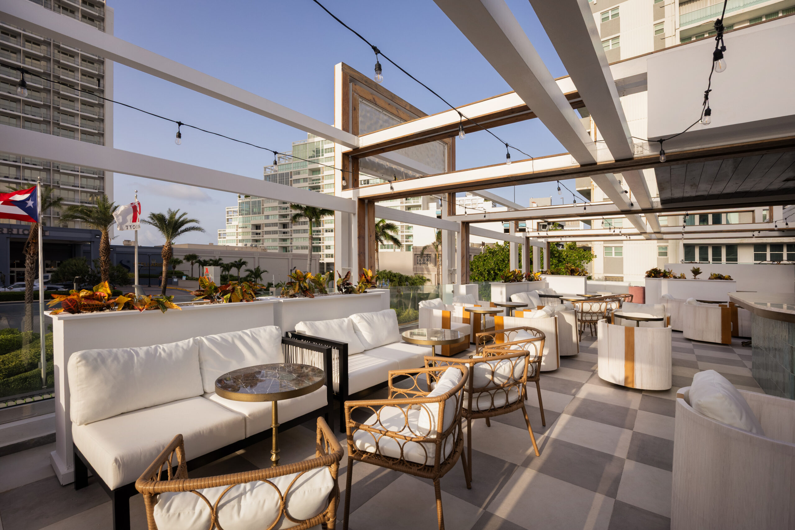 Rooftop patio with white cushioned seating, wicker chairs, small tables, and string lights, overlooking modern buildings on a sunny day.